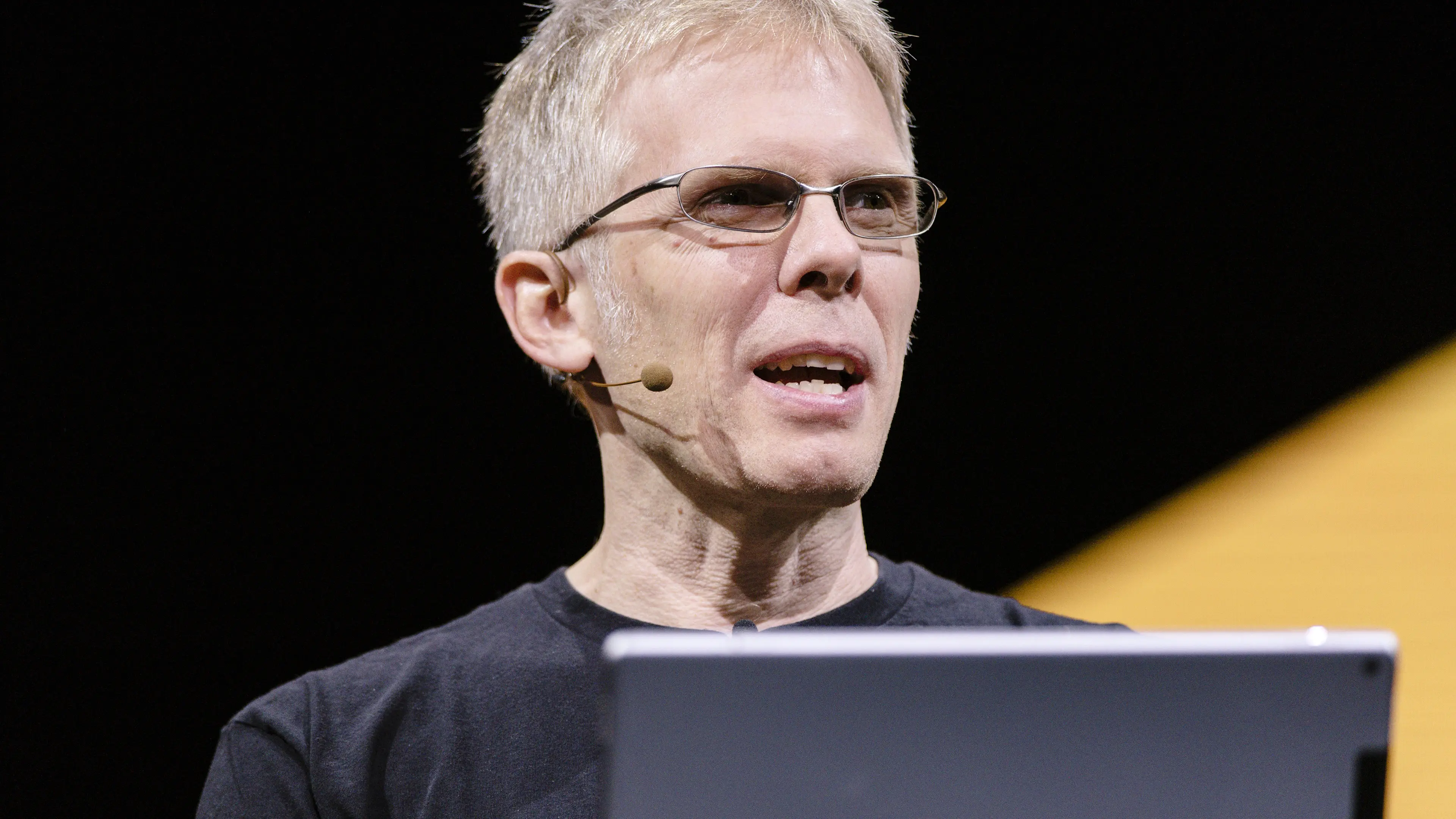 John Carmack speaking at a conference with a microphone and laptop.